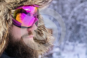 Close-up portrait of an attractive bearded man in a ski mask and a fur hat against the background of a snowy forest
