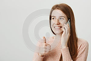 Close-up portrait of attractive ginger girl with cute freckles speaking on cellphone while showing thumbs up and smiling