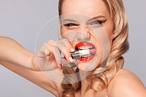Close up portrait of attractive girl rouging her lips. She is holding red lipstick in the mouse. Isolated on gray
