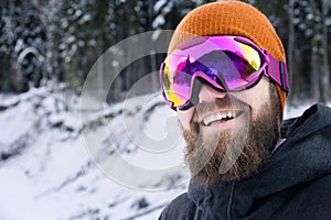 Close-up portrait of an attractive laughing young bearded man in a knitted winter hat and ski mask with goggles on his
