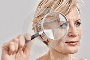 Close up portrait of attractive middle aged woman looking aside, holding a magnifying glass and showing her wrinkles