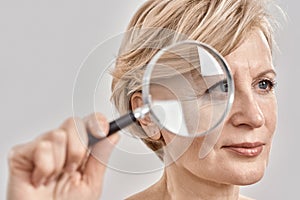Close up portrait of attractive middle aged woman looking aside, holding a magnifying glass and showing her wrinkles