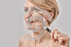 Close up portrait of attractive middle aged woman looking aside, holding a magnifying glass and showing her wrinkles