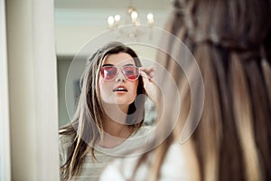 Close-up portrait of attractive modern european woman in optician store standing near mirror and trying on stylish