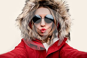 Close up portrait of attractive woman blowing red lips sending sweet air kiss stretching hand for taking selfie wearing red jacket