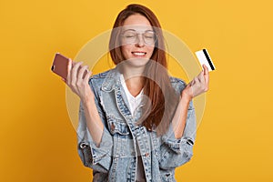 Close up portrait of attractive woman holding cell phone and credit card in hands, keeps eyes closed, looks happy, her dream cames