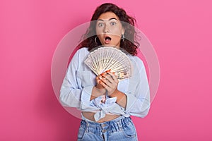 Close up portrait of attractive woman holding fan of money in her hands, keeps mouth opened, wearing stylish clothng, wins lottery photo