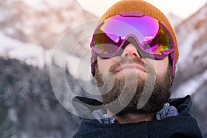 Close-up portrait of an attractive young bearded man in a knitted winter hat and ski mask with goggles on his face in