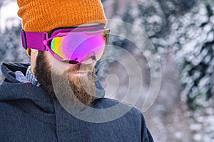 Close-up portrait of an attractive young bearded man in a knitted winter hat and ski mask with goggles on his face in