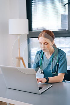 Close-up portrait of attractive young female practitioner doctor wearing blue green medical uniform working typing on