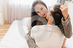 Close-up portrait from back of brunette guy embracing pretty european girl in bedroom. Indoor photo of smiling beautiful