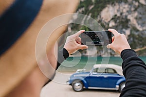 Close-up portrait from back of guy in light-brown hat taking photo of vintage car standing on mountain background