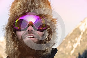 Close-up portrait of a bearded happy snowboarder skier in a ski mask with goggles and a fur big old-school hat on a