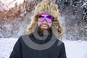 Close-up portrait of a bearded happy snowboarder skier in a ski mask with goggles and a fur big old-school hat on a