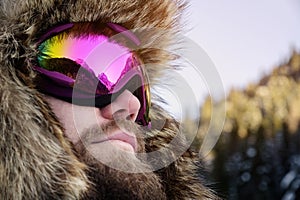 Close-up portrait of a bearded happy snowboarder skier in a ski mask with goggles and a fur big old-school hat on a