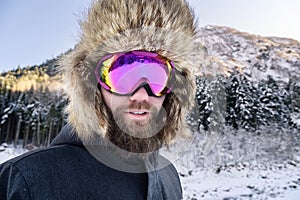 Close-up portrait of a bearded happy snowboarder skier in a ski mask with goggles and a fur big old-school hat on a