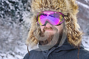 Close-up portrait of a bearded happy snowboarder skier in a ski mask with goggles and a fur big old-school hat on a