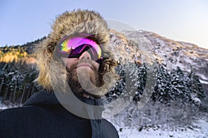 Close-up portrait of a bearded happy snowboarder skier in a ski mask with goggles and a fur big old-school hat on a