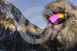 Close-up portrait of a bearded happy snowboarder skier in a ski mask with goggles and a fur big old-school hat on a