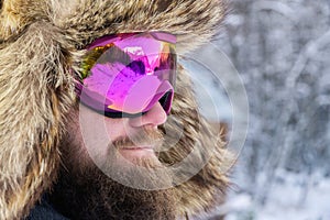 Close-up portrait of a bearded happy snowboarder skier in a ski mask with goggles and a fur big old-school hat on a