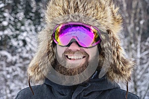 Close-up portrait of a bearded happy snowboarder skier in a ski mask with goggles and a fur big old-school hat on a