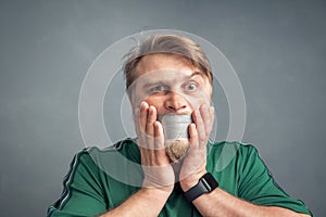 A close-up portrait of a bearded man with his mouth taped shut, holding his hands over his face in horror