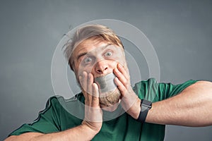 A close-up portrait of a bearded man with his mouth taped shut, holding his hands over his face in horror