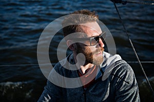 Close up portrait Bearded man model wearing sunglasses and hoodie looks away, sunset in the sea scenery.