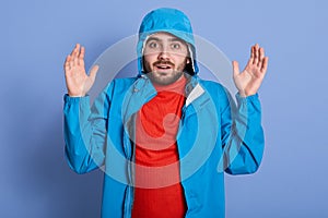 Close up portrait of bearded man standing with hands up wearing blue jacket with hood and red shirt, posing isolated over studio