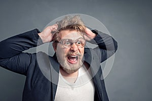 A close-up portrait of a bearded young man with a shocked face and tousled hair in huge glasses grabs his head with his