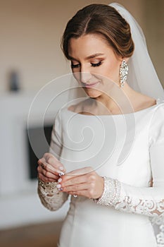 Close up portrait of beautiful bride standing by the window at home and tying on earrings. Charming bride in white