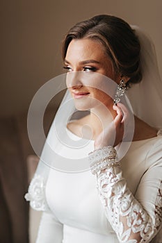 Close up portrait of beautiful bride standing by the window at home and tying on earrings. Charming bride in white