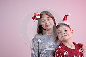 Close up portrait of beautiful children in santa sweaters and hats isolated over pink background, sister hugging her