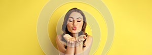 Close up portrait of beautiful, coquettish woman sending air kiss, blowing mwah at camera, standing over yellow