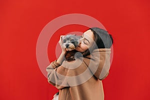 Close up portrait of a beautiful dog-owner female in a hoodie hugging her adorable york terrier in front of the  red wall