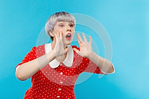 Close up portrait of beautiful dollish girl with short light violet hair wearing red dress shouting over blue background