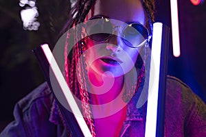 Close up portrait of a beautiful girl with an cornrows, wearing a denim jacket and sunglasses, holds in her hands neon night club