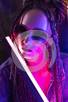 Close up portrait of a beautiful girl with an cornrows, wearing a denim jacket and sunglasses, holds in her hands neon night club