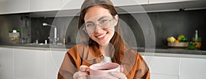 Close up portrait of beautiful girl in glasses, holding hot cup of drink, drinking tea or coffee at home and smiling at