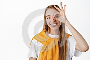 Close up portrait of beautiful happy girl with white smile, showing OK okay gesture near eye, approve something good