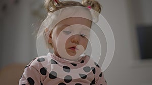 Close-up portrait of beautiful little girl with ponytails looking down. Sad pretty cute Caucasian blond baby indoors at