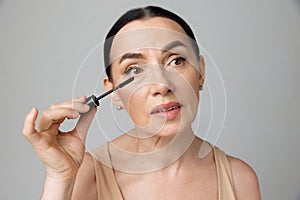 Close up portrait of beautiful mature brunette woman applying mascara with makeup brush over grey background. Fashion