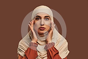 Close-up portrait of beautiful modern muslim woman with natural make-up dressed in beige hijab posing on brown background in