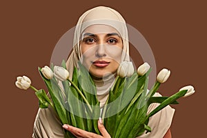 Close-up portrait of a beautiful Muslim woman dressed in a beige hijab who looks at the camera and holds a large bouquet of tulips