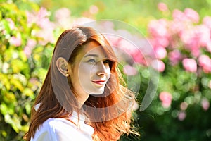 Close up portrait of a beautiful red haired girl in spring park. Pretty model between pink roses. Summer freedom enjoy