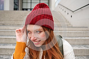 Close up portrait of beautiful redhead girl in red hat, urban woman with freckles and ginger hair, sits on stairs on