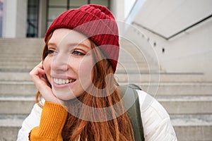 Close up portrait of beautiful redhead girl in red hat, urban woman with freckles and ginger hair, sits on stairs on