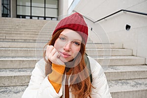 Close up portrait of beautiful redhead girl in red hat, urban woman with freckles and ginger hair, sits on stairs on