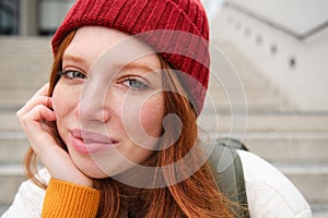 Close up portrait of beautiful redhead girl in red hat, urban woman with freckles and ginger hair, sits on stairs on