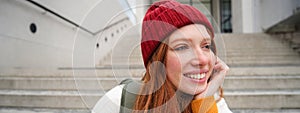 Close up portrait of beautiful redhead girl in red hat, urban woman with freckles and ginger hair, sits on stairs on